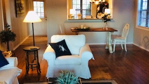 Ein Wohnzimmer mit weißem Sessel, blauer Kommode, Stehlampe und Essbereich mit Tisch und Stühlen.