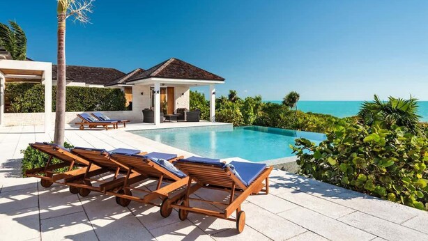 A poolside area with lounge chairs, a covered seating area, and a view of the ocean. - Beachfront Villa w/ Infinity Pool & Ocean Views (Long Bay)