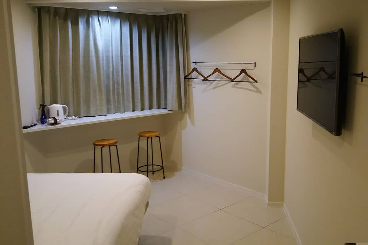 Een kleine hotelkamer met een bed, een raam met gordijnen, een aan de muur bevestigde televisie en twee krukjes.