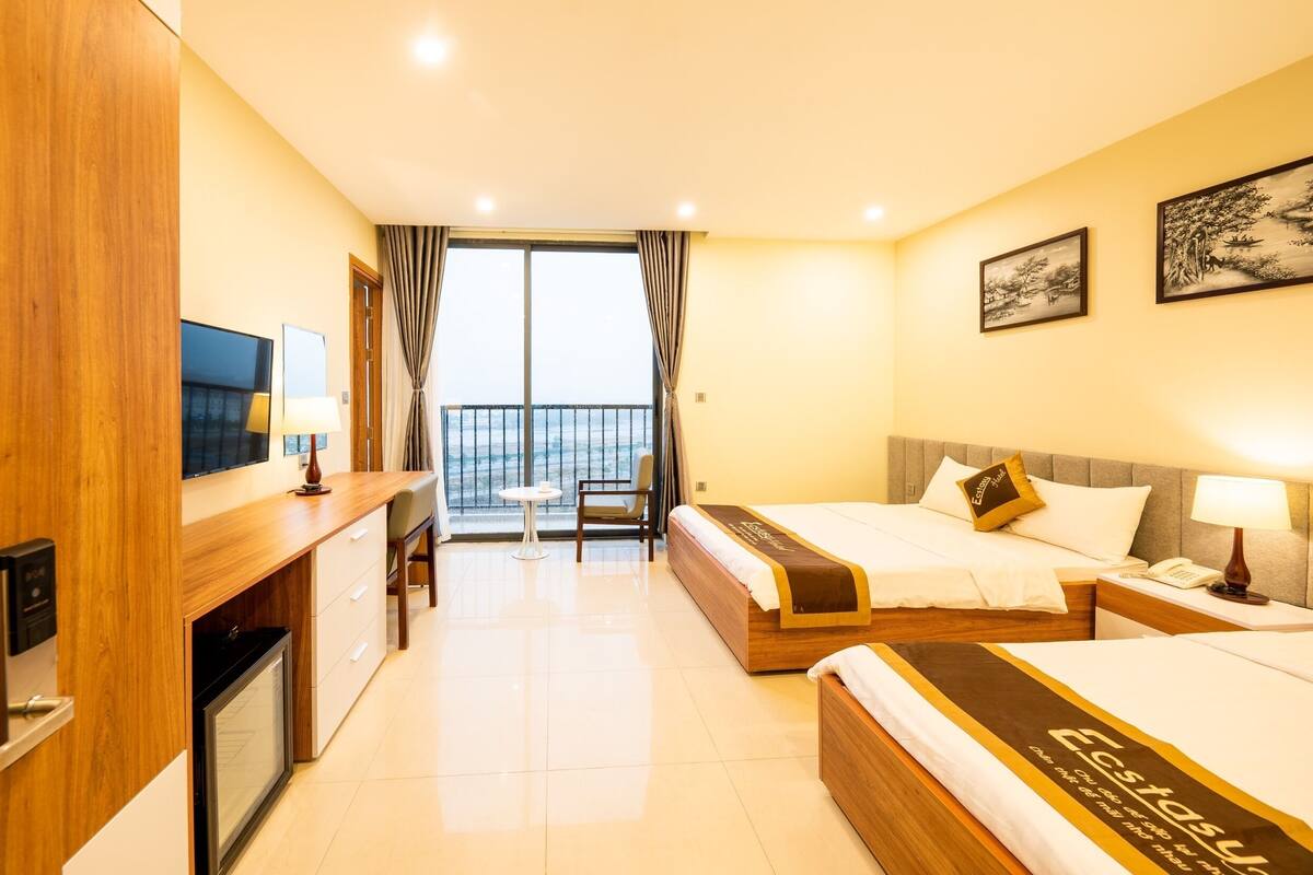 A hotel room with two beds, a desk, a TV, a balcony with curtains, and artwork on the walls.