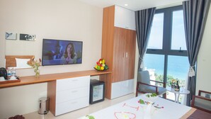 A modern hotel room with a large TV, a wooden desk, a mini-fridge, a small table with a bottle and glass, and a view of the sea through large windows. - Ecstasy Hotel (Nha Trang)