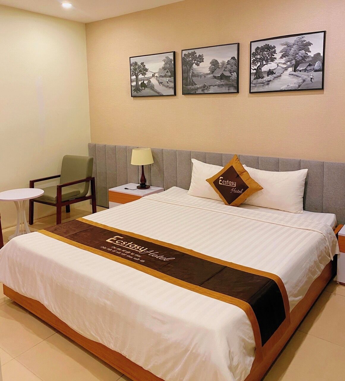 A hotel room with a bed, a chair, a small table, and three framed pictures on the wall.