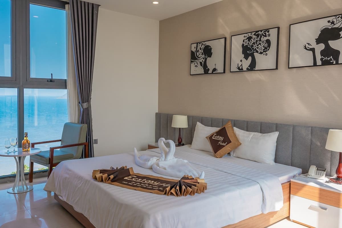 A hotel room with a bed, a bedside table, a lamp, a phone, and a view of the ocean.