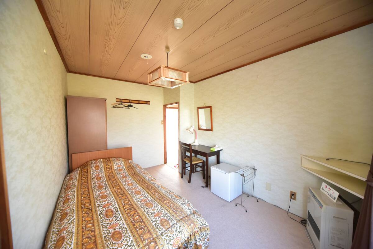 A bedroom with a bed, a desk, a chair, a small refrigerator, and a heater.