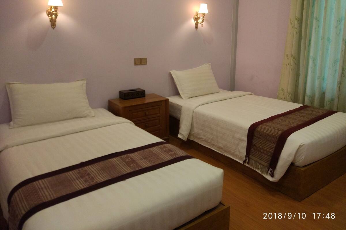 Une chambre d’hôtel avec deux lits simples, une table de chevet en bois, une lampe et un rideau.