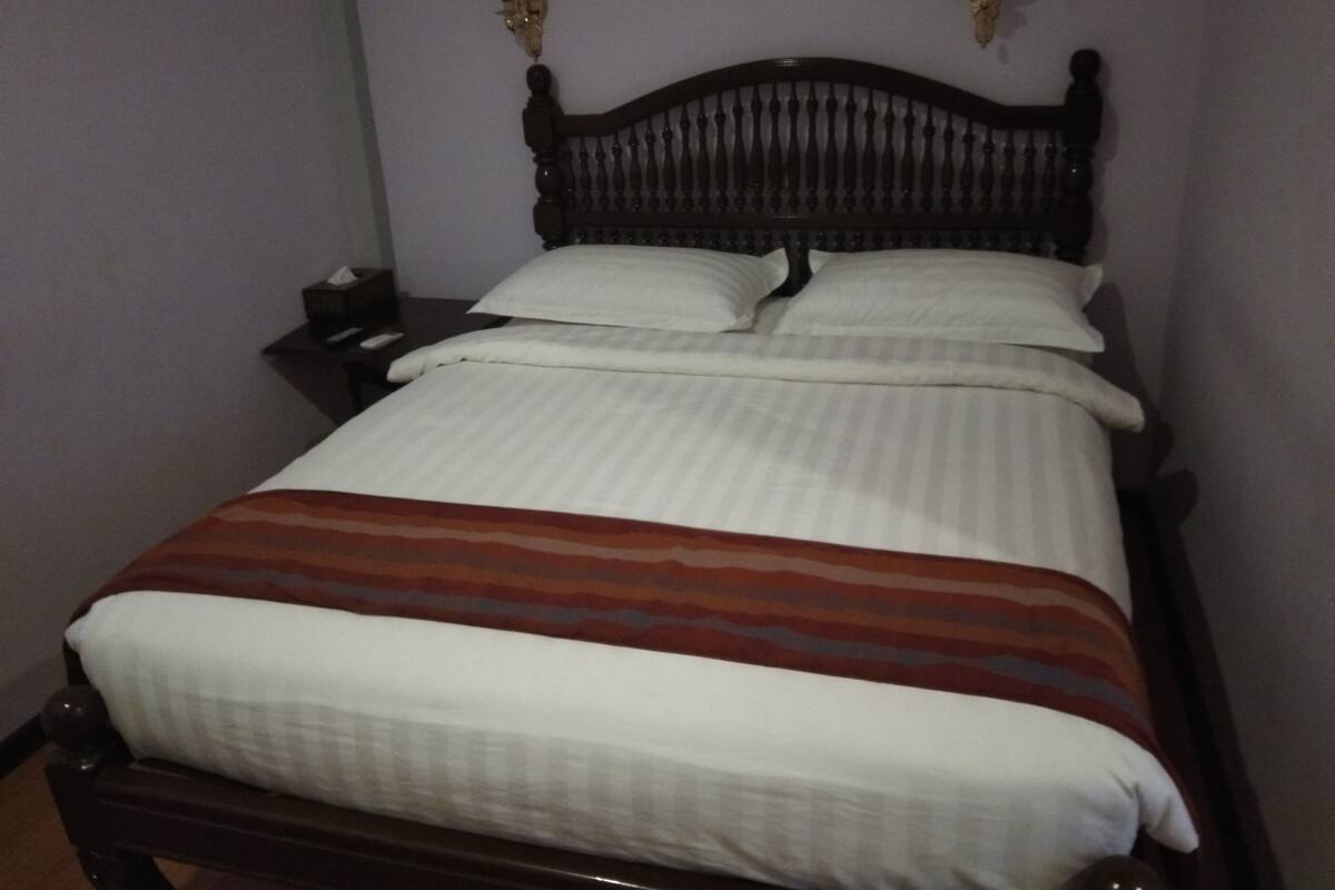 Un lit bien fait, avec une couverture à rayures, une tête de lit en bois et une table de chevet sur laquelle se trouve une lampe.