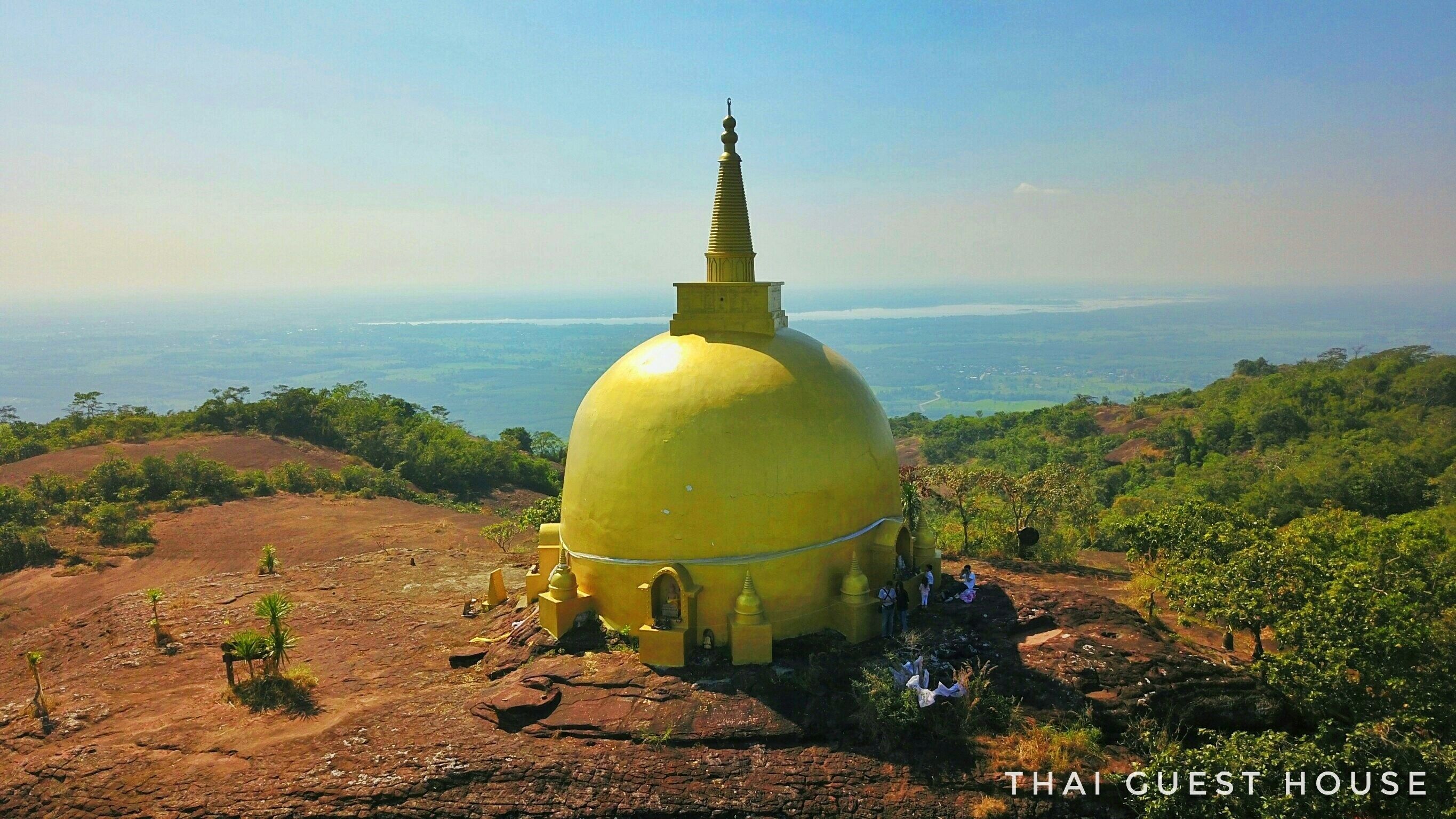 A large yellow stupa surrounded by rocky terrain and greenery, with a view of distant landscapes.