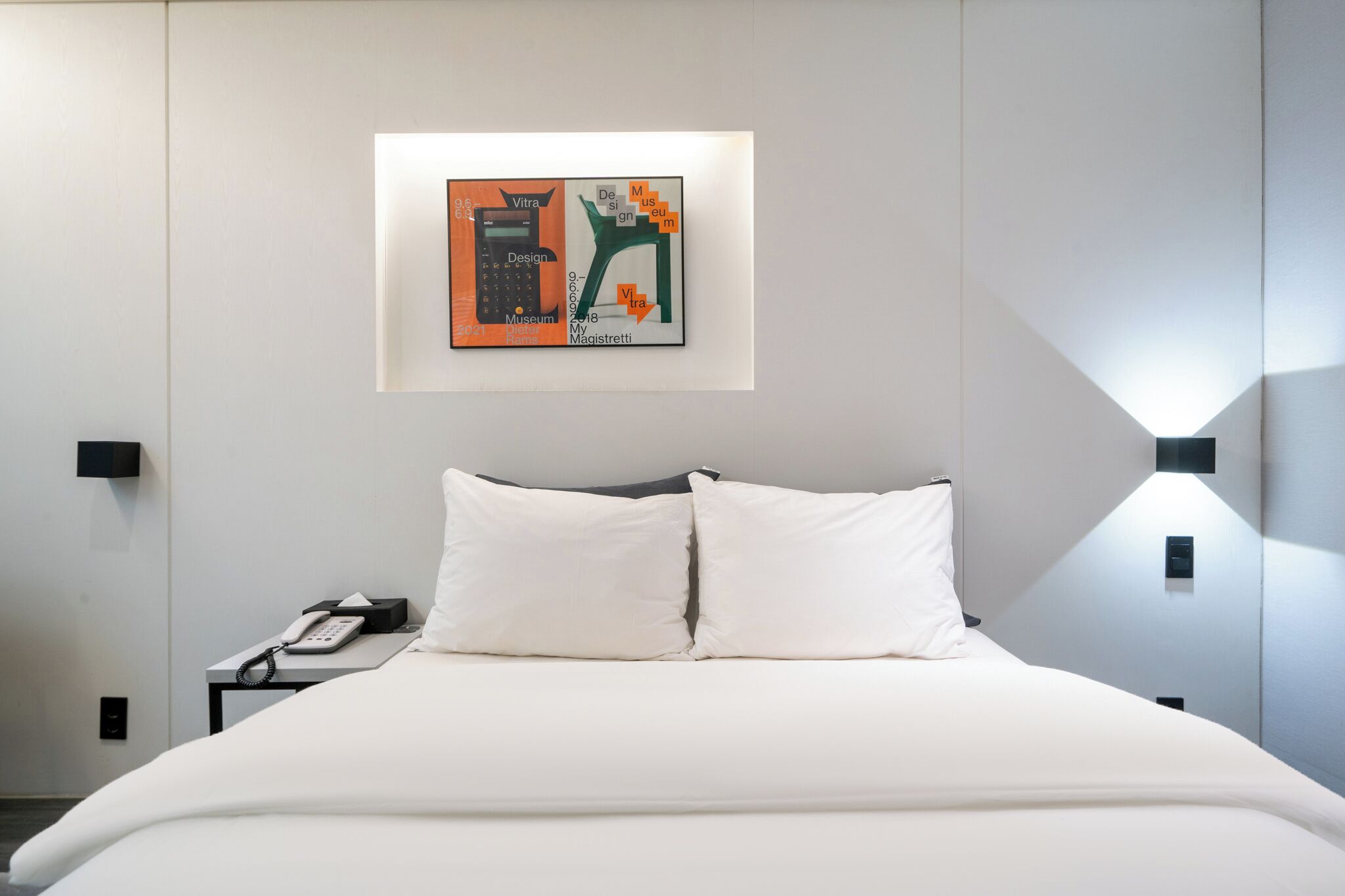 A hotel room with a bed, white pillows, a bedside table with a phone, and a wall-mounted artwork.