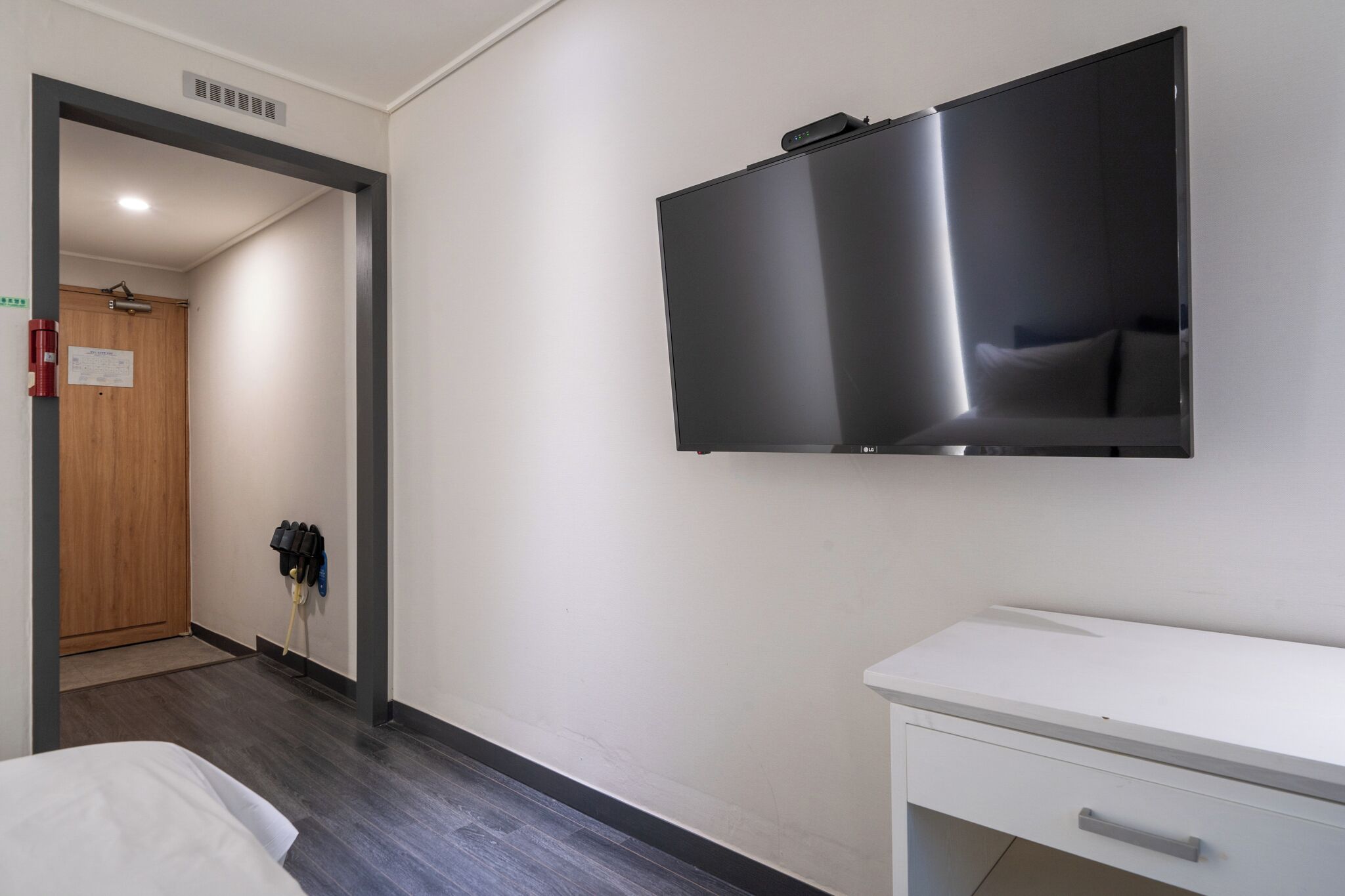 Une chambre d’hôtel avec un lit, une commode blanche, un téléviseur à écran plat fixé au mur, une porte en bois et un porte-manteau avec des vestes.