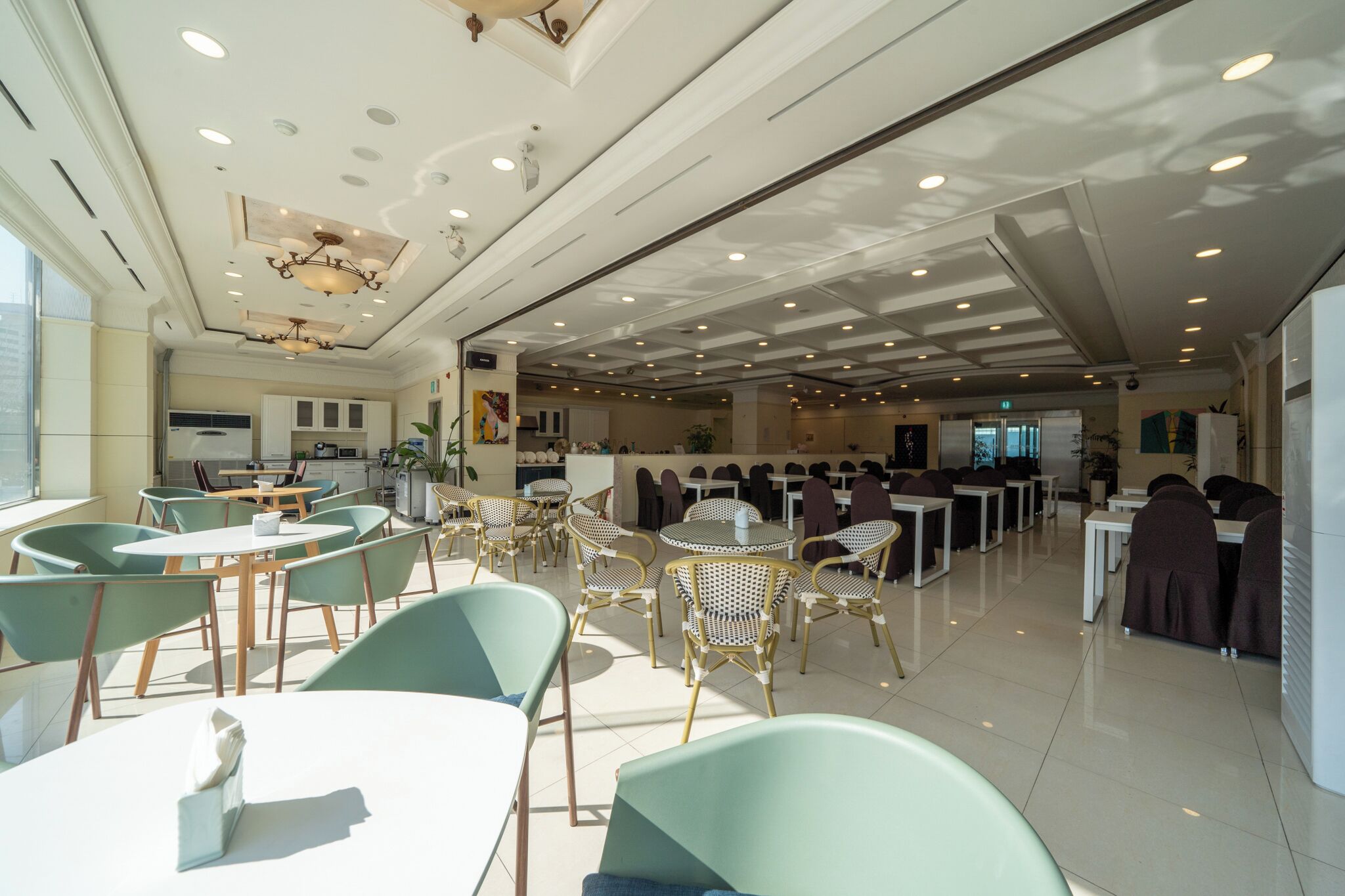 A spacious indoor dining area with multiple tables and chairs, a kitchen in the background, and a modern ceiling design.