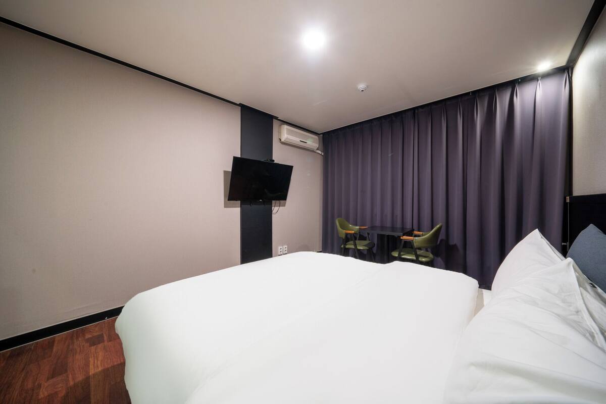 A hotel room with a large bed, a flat-screen TV mounted on the wall, a small table with two chairs, and a window with dark curtains.