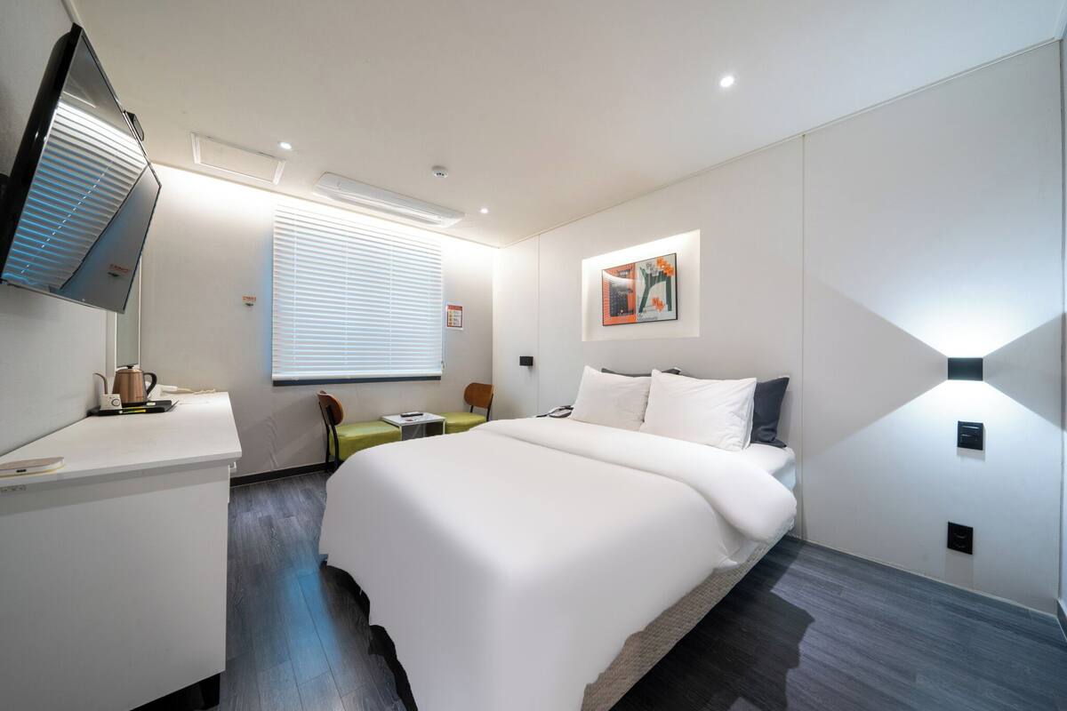 A modern hotel room with a large bed, a desk with a kettle, a chair, a bedside table, a flat-screen TV mounted on the wall, and a window with blinds.