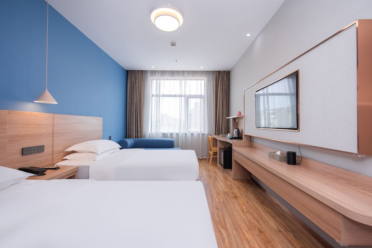A hotel room with two beds, a desk, a chair, a TV, and a large window with curtains.