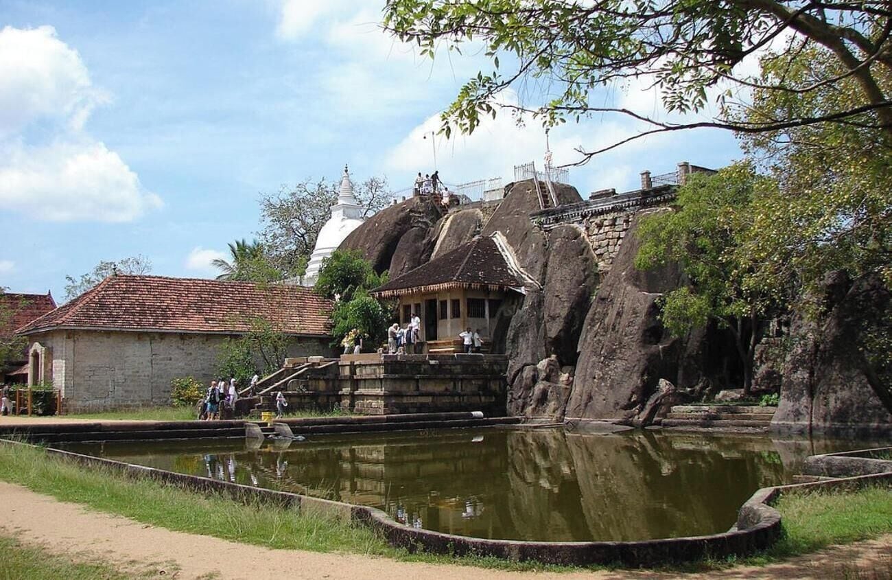 A traditional temple with a stone structure, a water body in front, and a white building in the background.