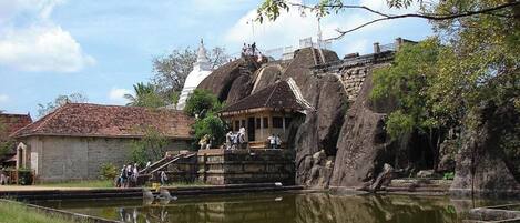 A traditional temple with a stone structure, a water body in front, and a white building in the background.