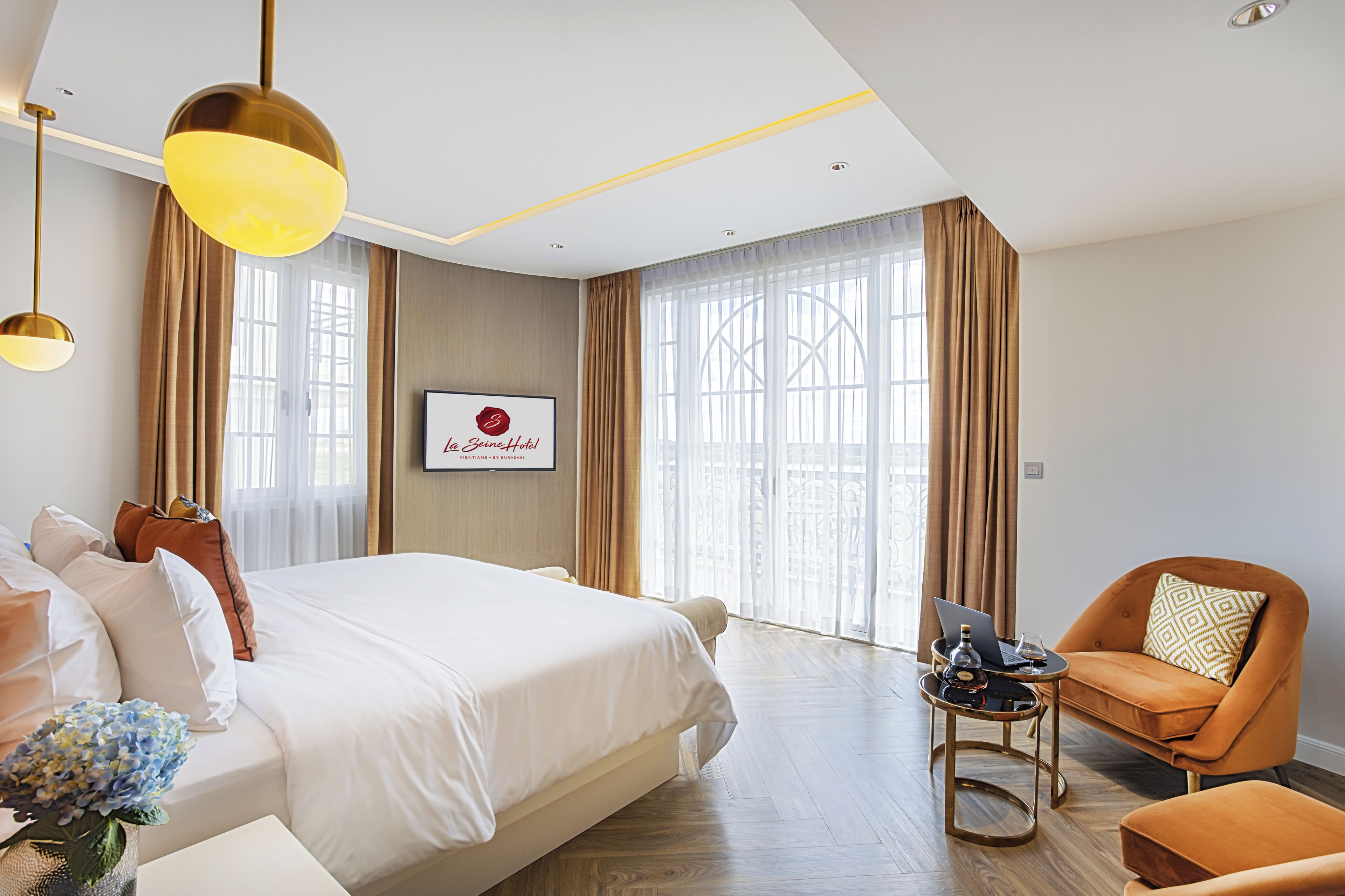 A hotel room with a large bed, a desk with a laptop, an orange armchair, and a large window with curtains.