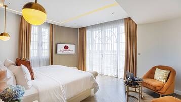 A hotel room with a large bed, a desk with a laptop, an orange armchair, and a large window with curtains.