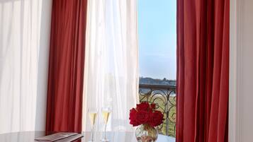 Una mesa con dos copas de champán y un florero de rosas rojas, frente a un fondo de cortinas rojas y blancas con vistas a un balcón.