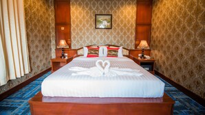 A large bed with a heart-shaped towel arrangement, wooden headboard, two bedside tables with lamps, and patterned wallpaper. - Soubandith Hotel (Pakse)