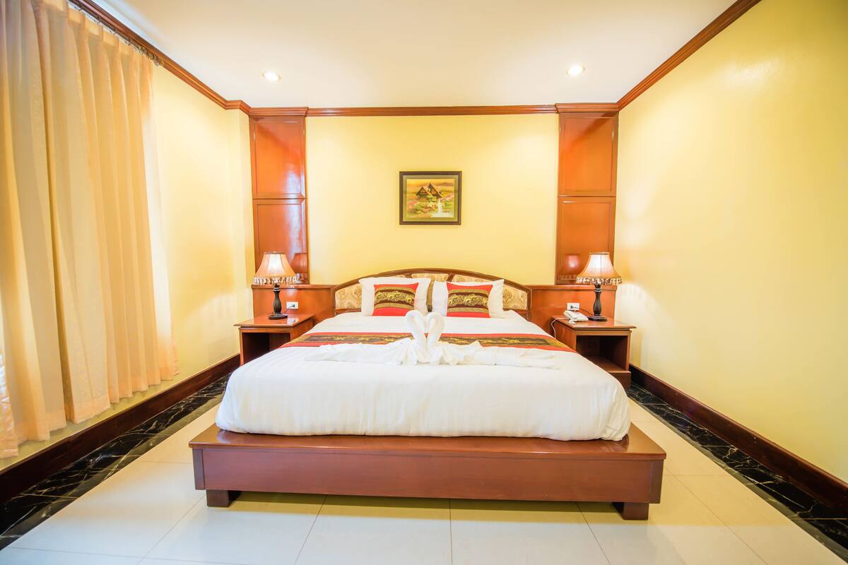 A large bed with a wooden frame, two bedside tables with lamps, a painting on the wall, and a curtain on the left.
