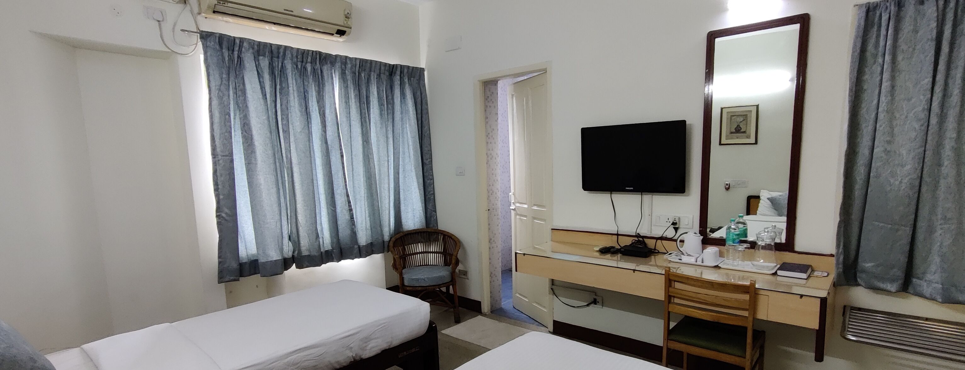 A hotel room with two single beds, a desk with a chair, a flat-screen TV, a mirror, and an air conditioning unit.