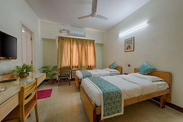 Une chambre d’hôtel avec deux lits simples, un bureau avec une chaise, une télévision, un climatiseur et un ventilateur de plafond.