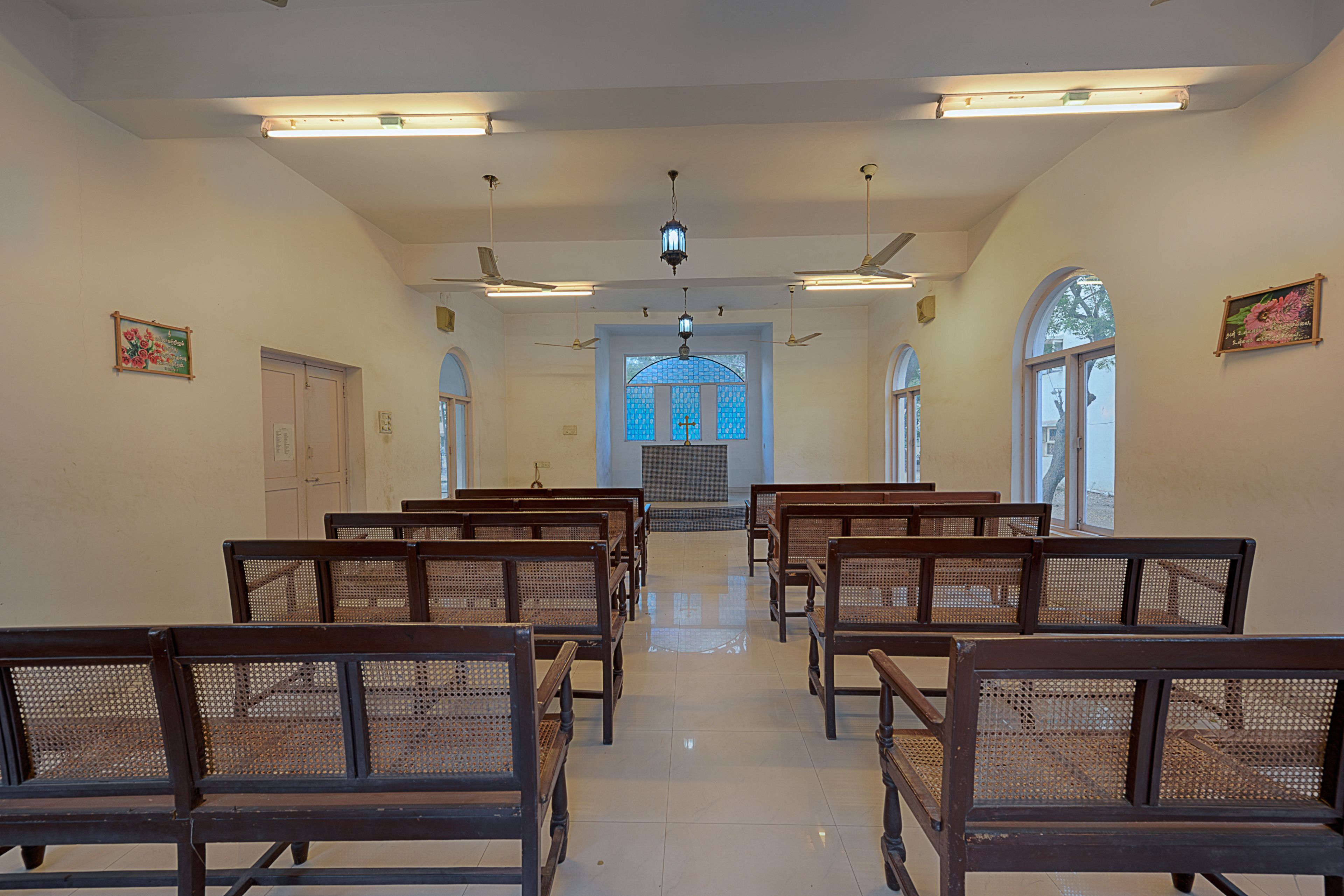 Ein Kircheninneres mit reihenweise angeordneten Kirchenbänken, einem Glasfenster und Deckenventilatoren.