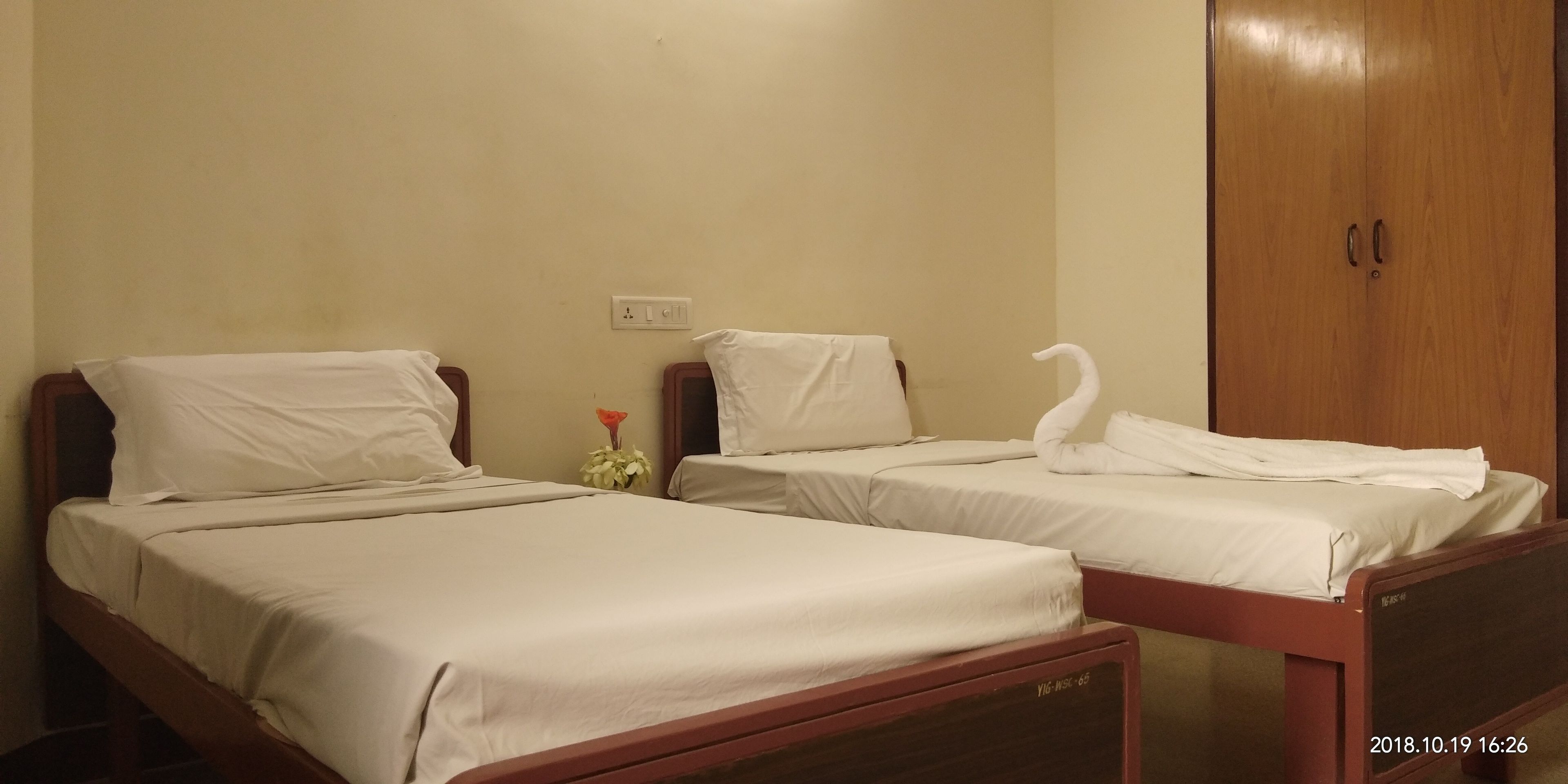 Two single beds with wooden frames and white bedding, a wooden wardrobe, and a small table with a red flower.