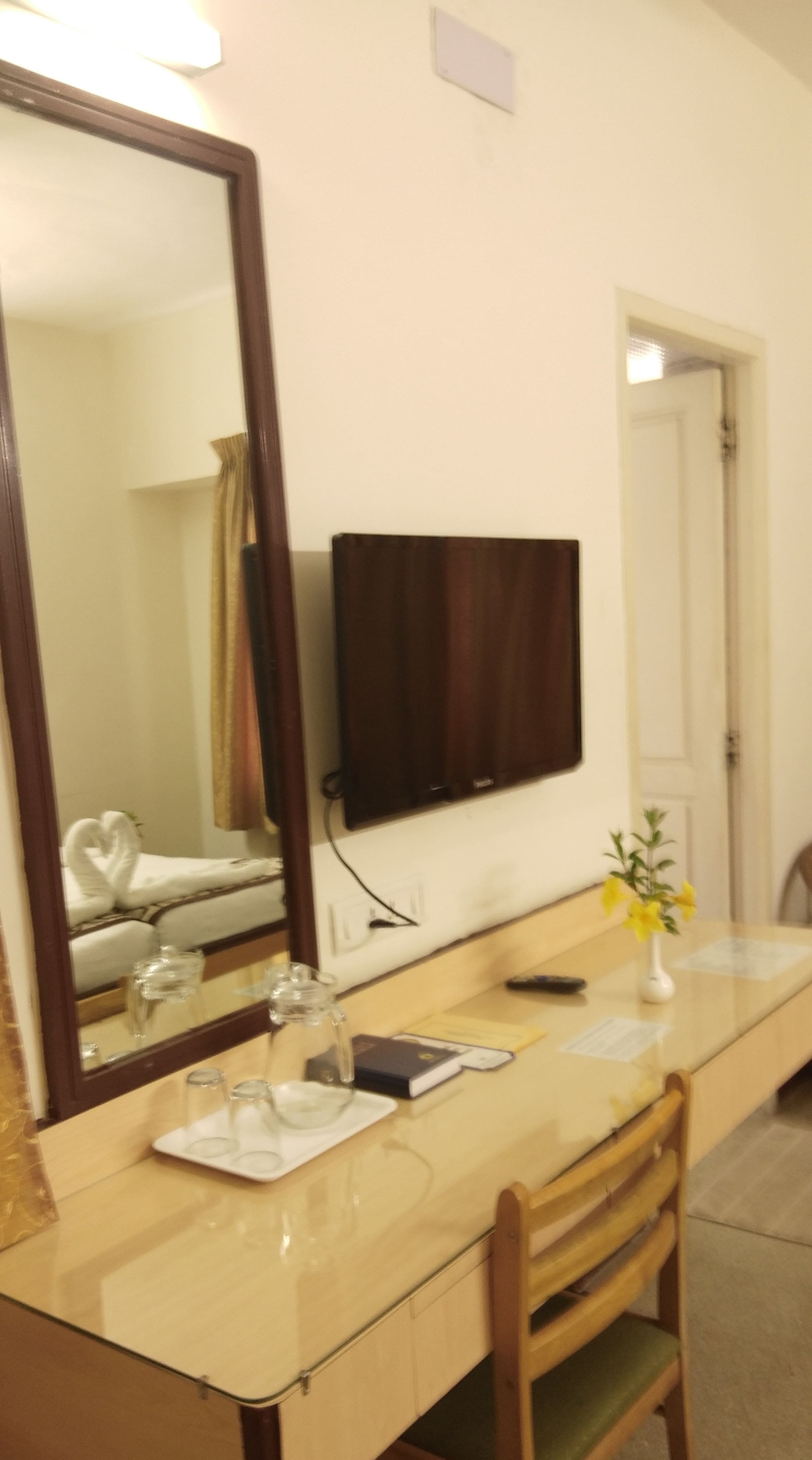 A hotel room with a large mirror, a flat-screen TV, a wooden chair, a glass table, and a vase with flowers.