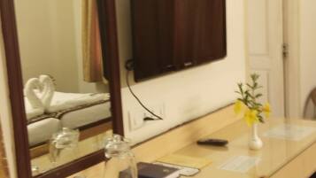 A hotel room with a large mirror, a flat-screen TV, a wooden chair, a glass table, and a vase with flowers.