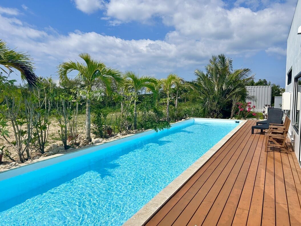 A swimming pool with a wooden deck, surrounded by palm trees and a building.