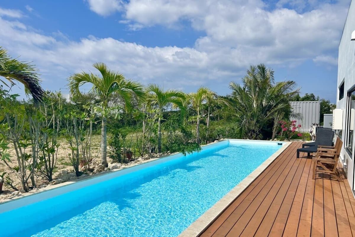 A swimming pool with a wooden deck, surrounded by palm trees and a building.