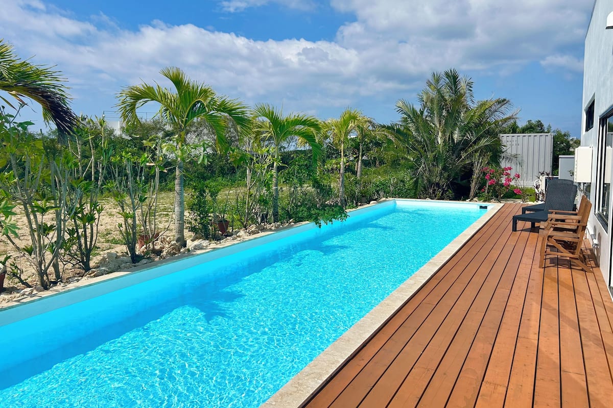 A rectangular swimming pool with a wooden deck, surrounded by tropical vegetation and a clear sky.