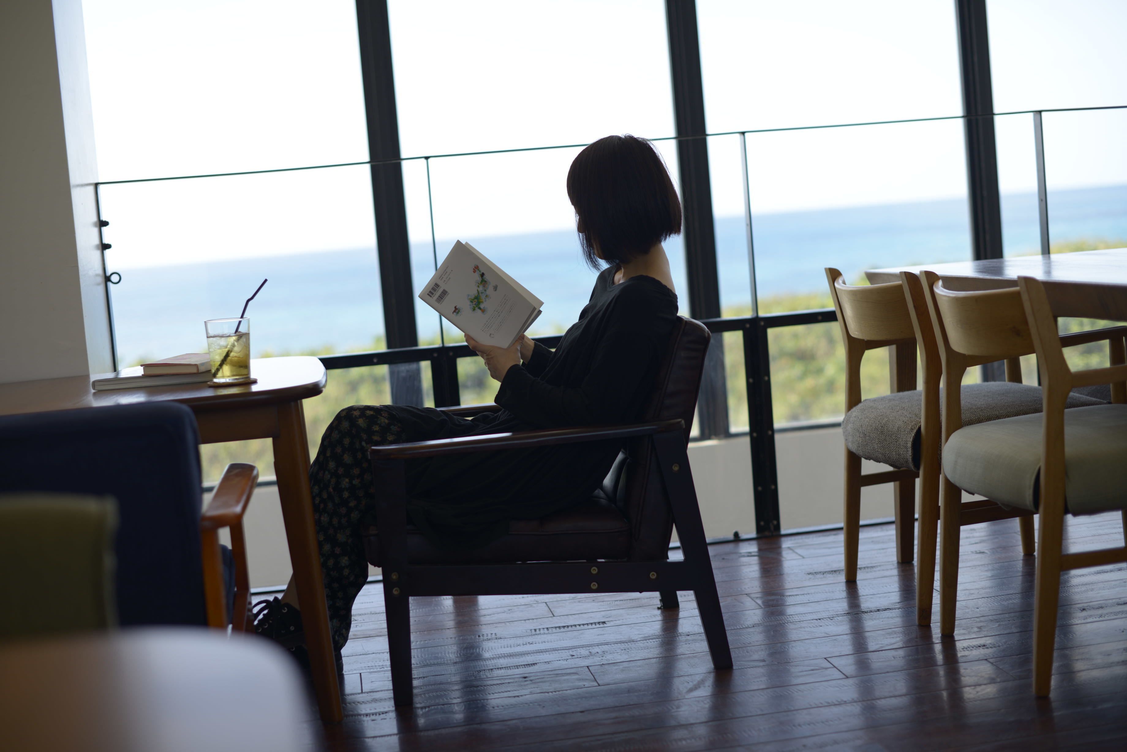 A person reading a book in a room with large windows overlooking the sea.