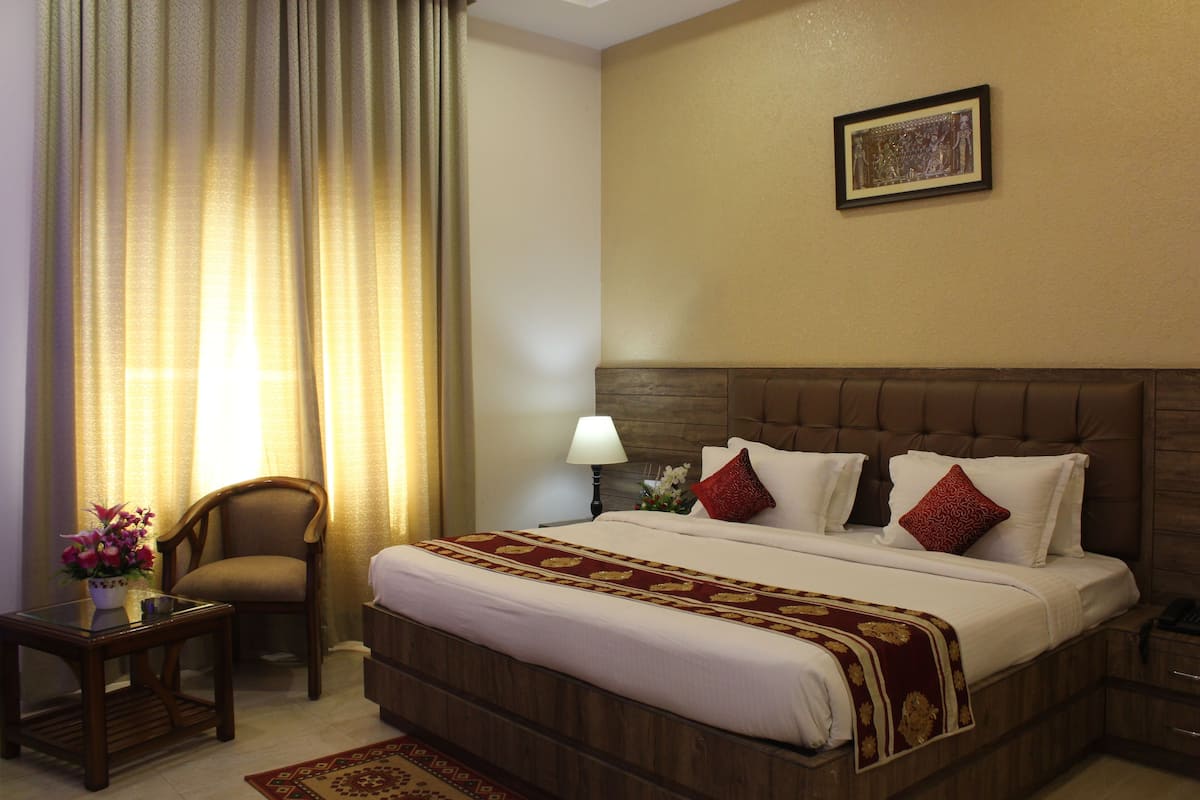 Habitación de hotel con una cama grande, cabecera de madera, mesita de noche, lámpara, silla y ventana con cortinas.