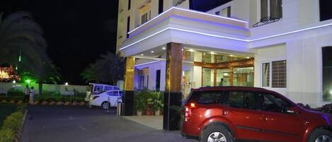 A hotel entrance with a red car parked in front, a white van, and a well-lit building facade.