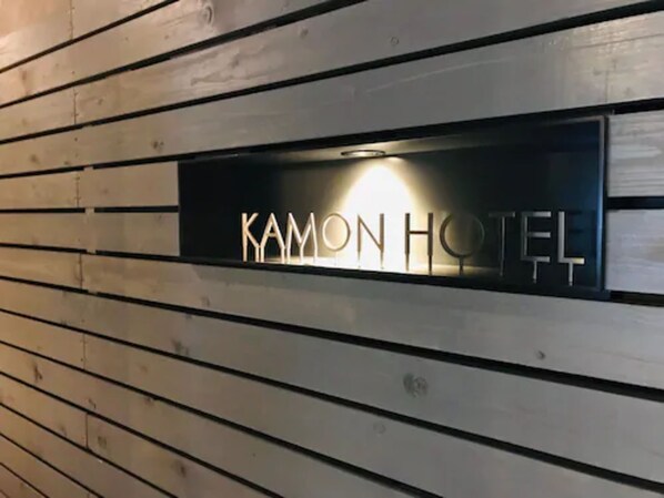 Une enseigne d’hôtel portant le nom « Kamon Hotel », fixée à un mur en bois.