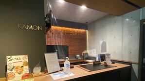 A reception desk with a wooden counter, a nameplate reading "KAMON", a menu stand, a bottle of hand sanitizer, and a small vase. - KAMON HOTEL Seto (Hiroshima)