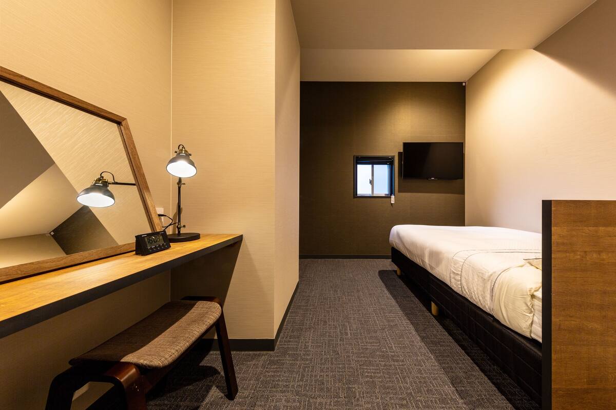 A hotel room with a wooden desk, bench, bedside table, bed, and a TV mounted on the wall.