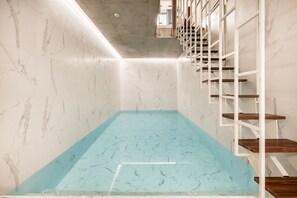 Un espace piscine intérieur moderne, avec une eau d’un bleu limpide, des murs en marbre et des étagères flottantes en bois.