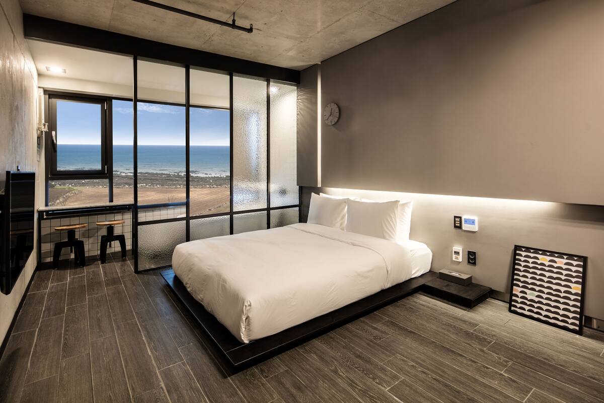 Une chambre moderne dotée d’une grande fenêtre donnant sur la plage, d’un lit avec du linge de lit blanc, d’une table de chevet et d’une horloge murale.