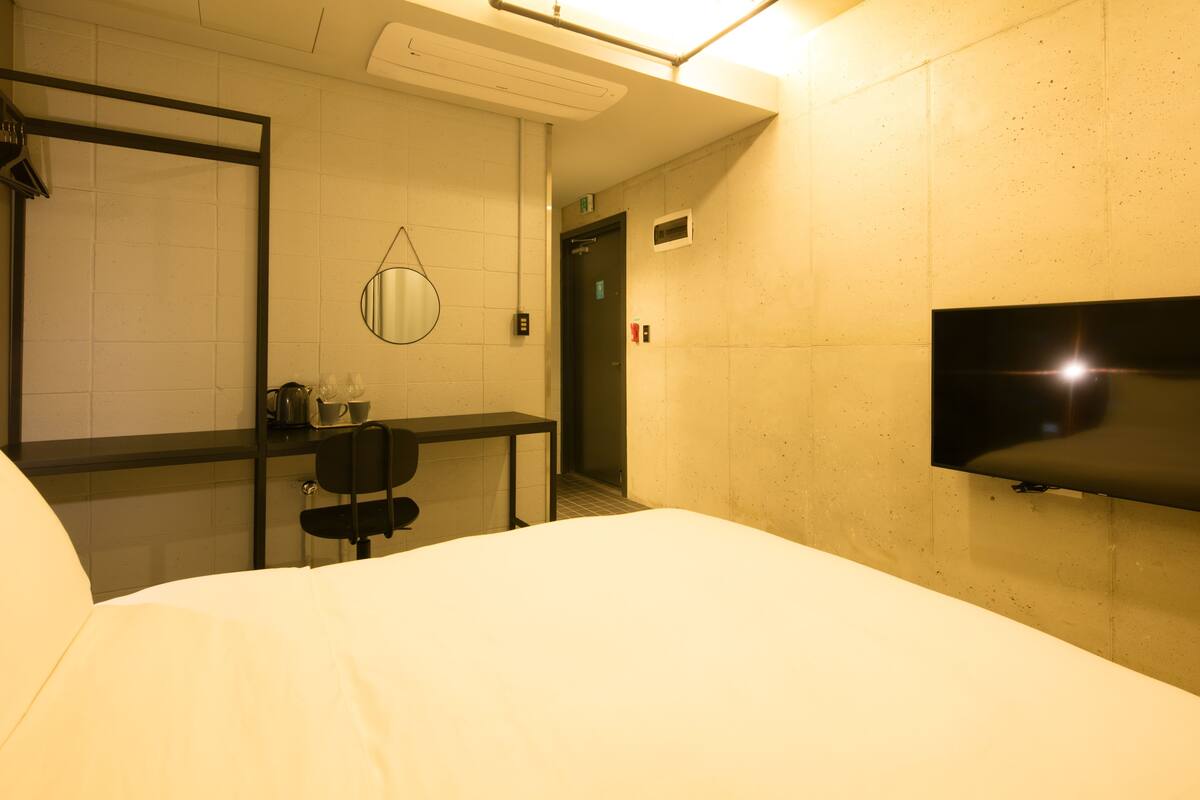 Une chambre d’hôtel comprenant un lit, un bureau avec une chaise, un miroir, une télévision et une porte donnant accès à un ascenseur.