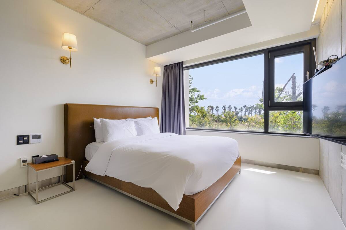 Une chambre d’hôtel moderne avec un grand lit, une table de chevet et une vue sur les palmiers, visible à travers une grande fenêtre.