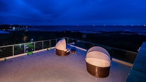 Terraza en la azotea con dos sillones grandes de mimbre y una mesa pequeña, con vistas a un paisaje costero de noche.