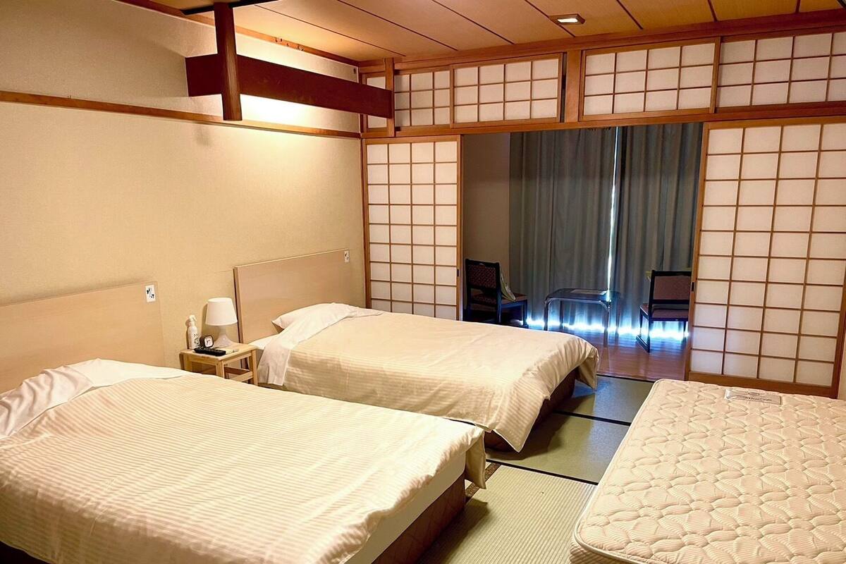 A traditional Japanese-style hotel room with two beds, tatami flooring, shoji screens, and a sliding door leading to a balcony.