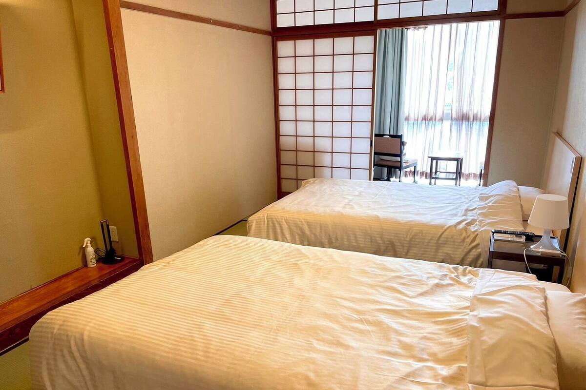 A hotel room with two beds, a wooden floor, a shoji screen, a desk with a lamp, and a view of the outdoors through a window with curtains.