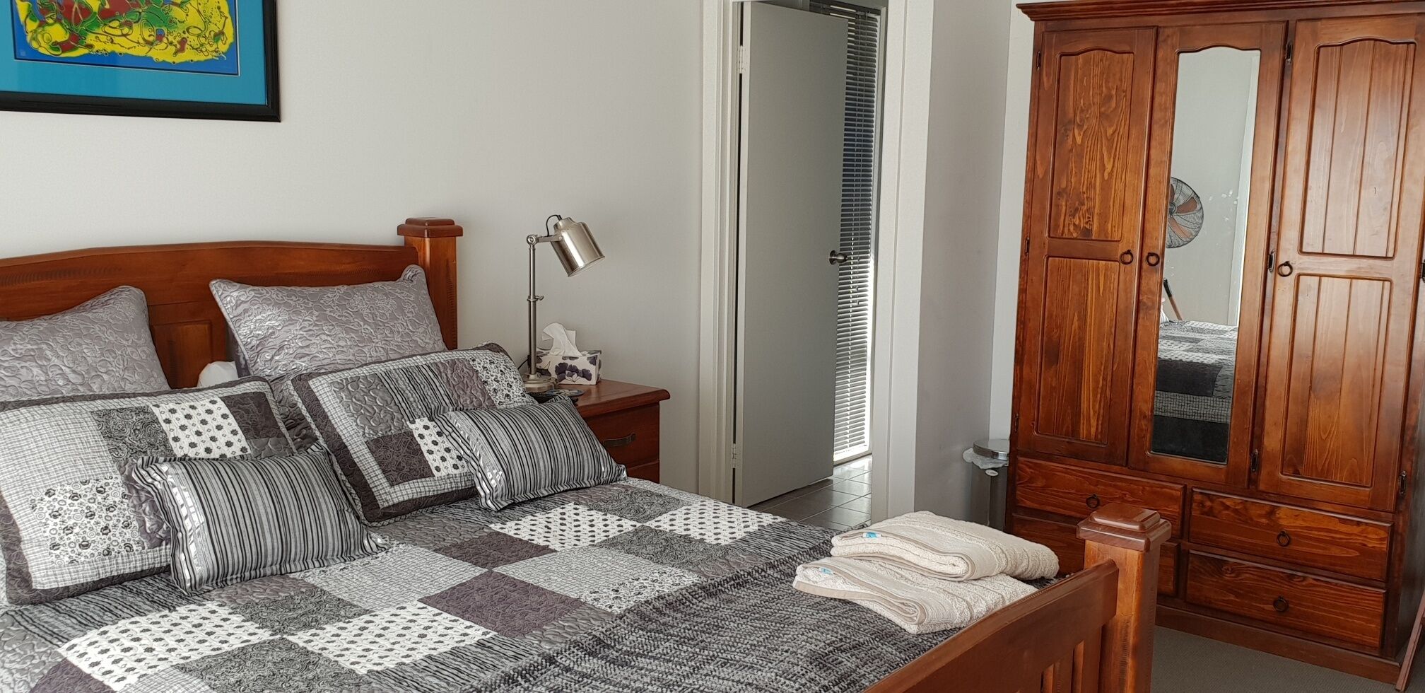 Une chambre à coucher avec un lit en bois, une table de chevet, une lampe, une armoire et une porte donnant sur une autre pièce.
