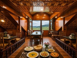 Un comedor con una mesa dispuesta para cuatro personas, rodeada de sillas de madera, con vista a un paisaje verde a través de grandes ventanales.