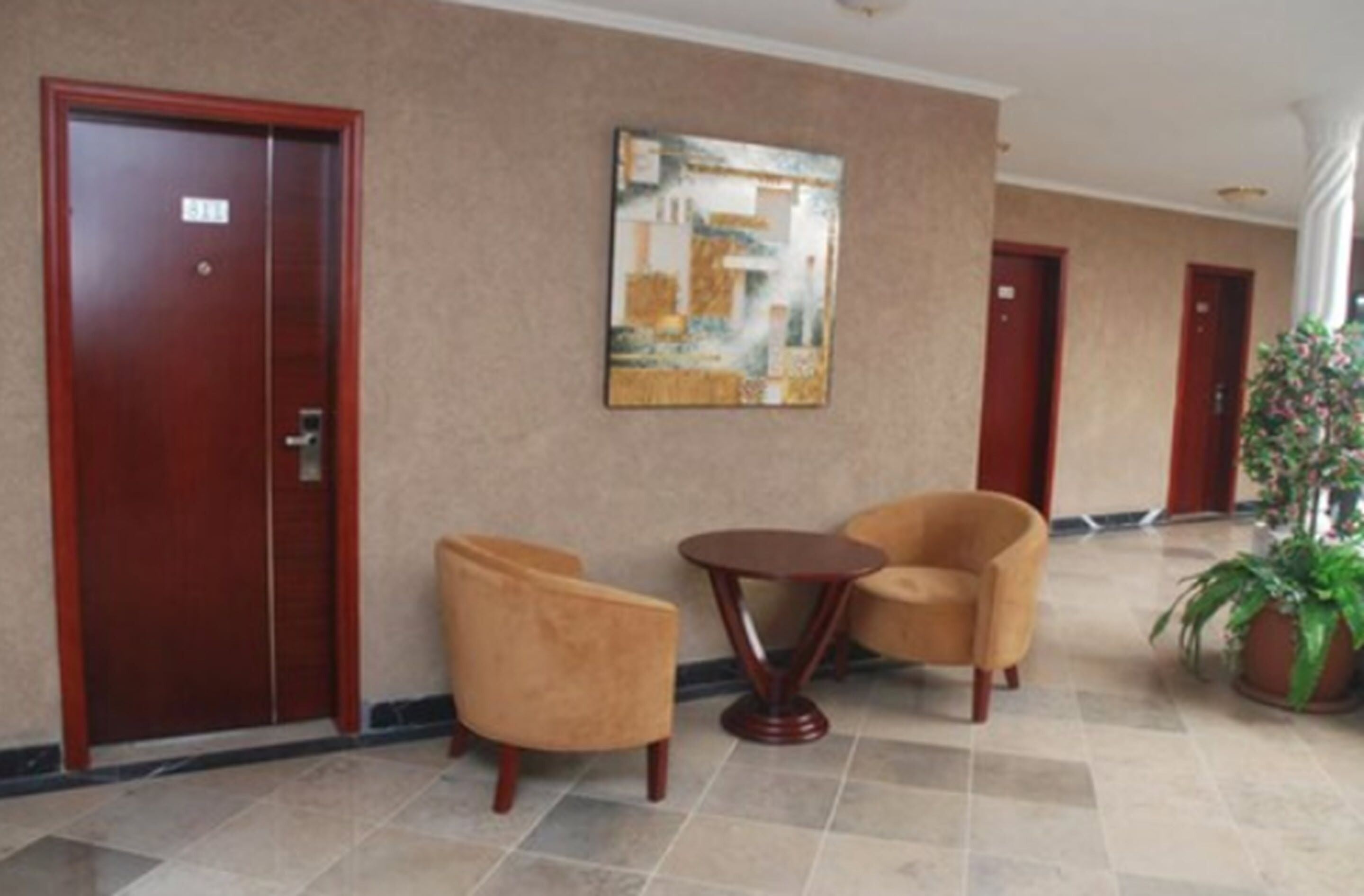 A lobby with a wooden door, a painting on the wall, two armchairs, a small round table, and a potted plant.