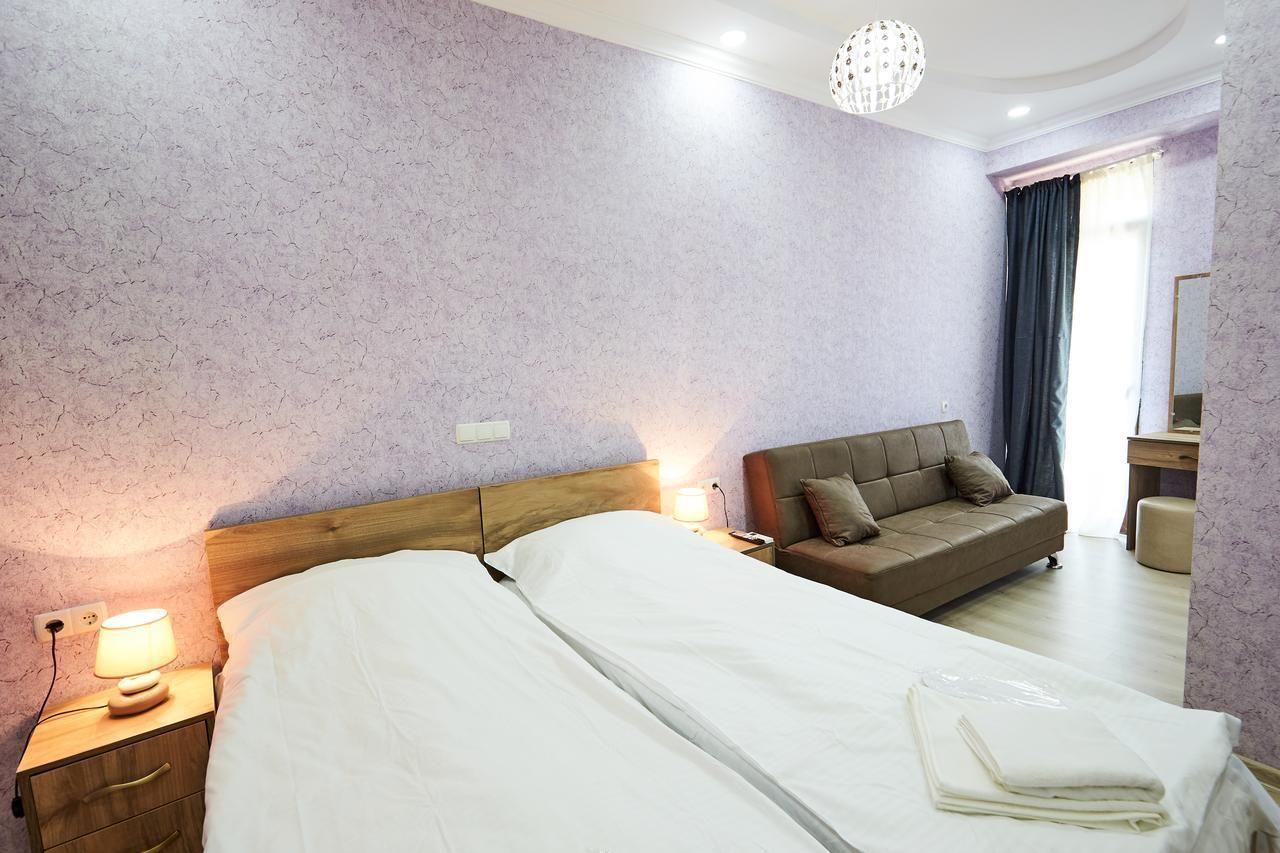A hotel room with a double bed, a sofa, a nightstand with a lamp, a mirror, and a view of the outside through a window.